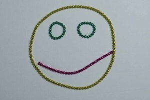 Gesicht aus Perlen geformt mit gelbem Kreis, zwei grünen Augen und einem schrägen lila Mund
