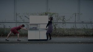 Zwei Frauen in Kleidern und hohen Schuhen bewegen einen weißen Küchenschrank auf einem Bürgersteig vor einer Hecke.