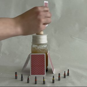 Hand baut Kartenhaus auf einem Glas mit Schnur, umgeben von zehn kleinen Schraubaufsätzen in einem Halbkreis.